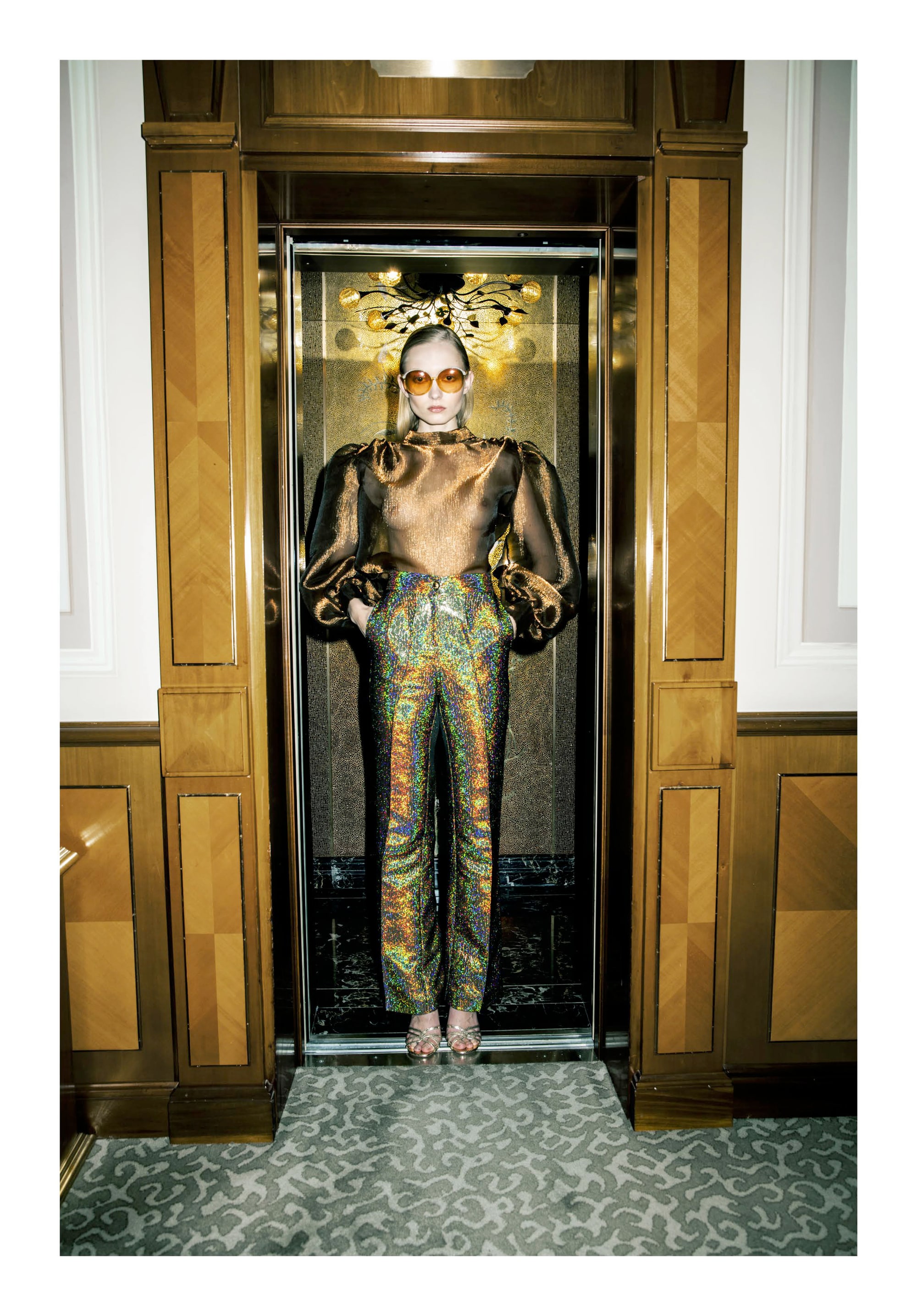 Nikita Lauffer in a bronze sheer blouse, glitter trousers and round orange sunglasses, standing in a lift